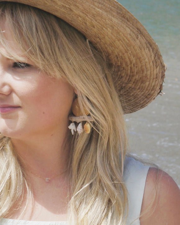 Gold, shell and driftwood earrings worn by woman with long blond hair in a straw hat on the beach