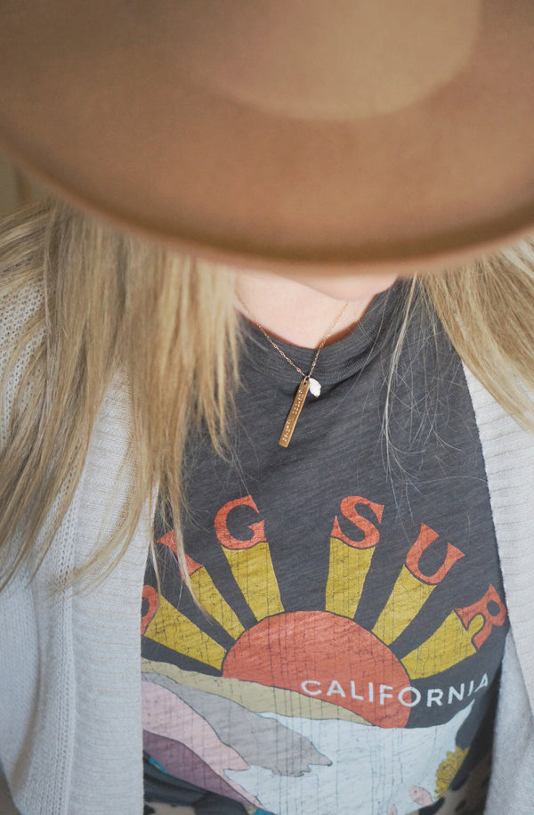 woman wearing happy place necklace with long blond hair, a hat, and a graphic tee