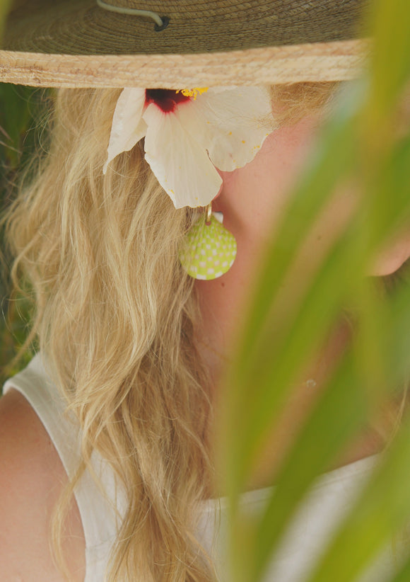 Lime green handpainted shell earrings in a checkered pattern worn by woman with long hair wearing a hat and flower in her hair