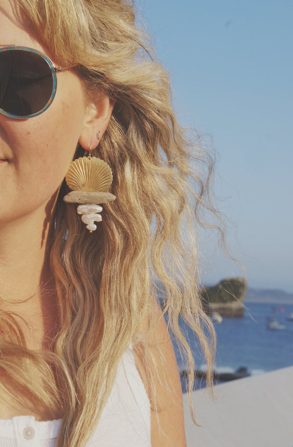 Gold shell, driftwood, and freshwater pearl earrings worn by woman with long blond hair in sunglasses with ocean in background